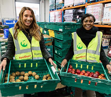 Two female volunteers in hi vis vests hold trays of surplus fruit.
