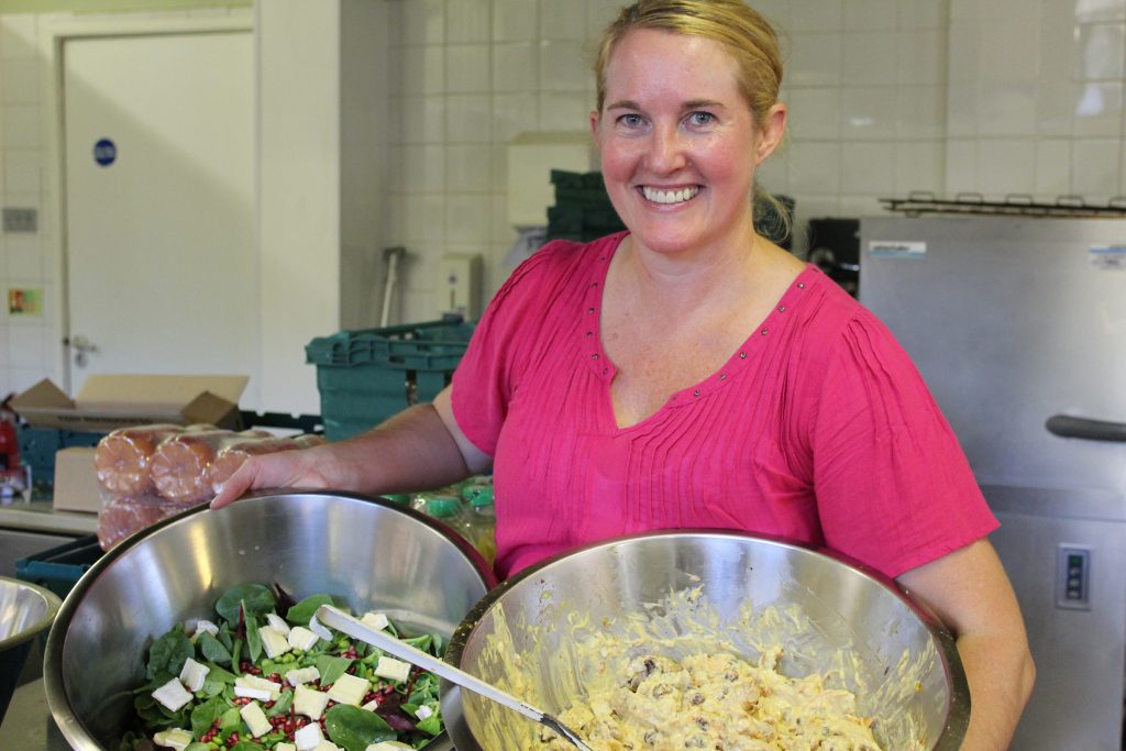 Claire, volunteer chef at Greenwich Migrant Hub, holds two cooking bowls of food as she smiles to the camera. One contains salad and one coronation chicken.