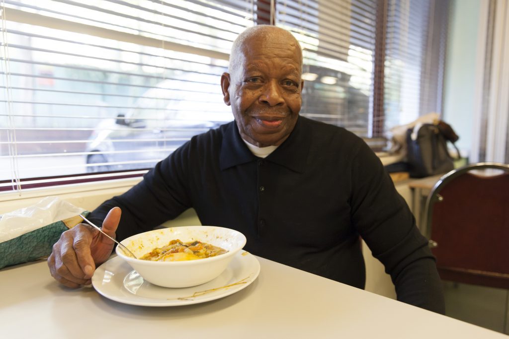 An older man smiles to the camera with a bowl of vegetable stew, made out of surplus food, in front of him. 