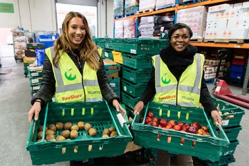 Two female volunteers in hi vis vests hold trays of surplus fruit.