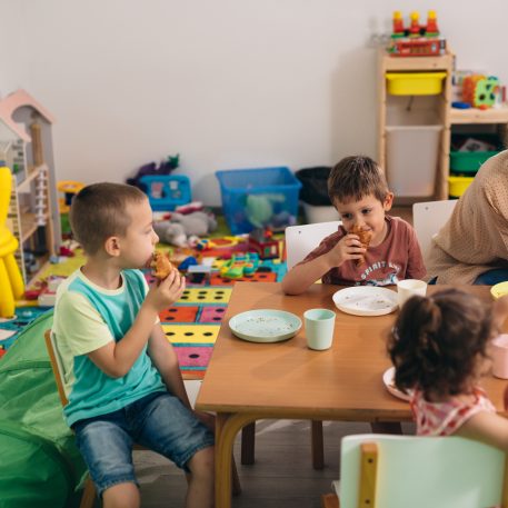 Children sitting round a table at a breakfast club, which has been set up at a nursery/primary school. A woman sits with them laughing and talking to them.